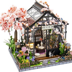 May include: A miniature greenhouse model featuring a black frame, a glass roof, and a cherry blossom tree with pink flowers. The structure includes a sign with Japanese text, a small cat figure, interior furniture, and a stone path in the garden.