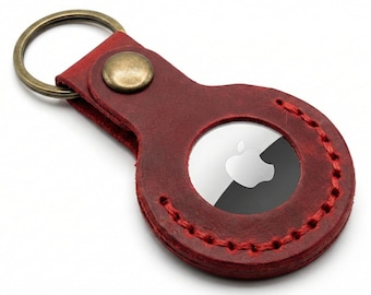 May include: A deep red leather keychain holder featuring a circular cutout that secures a silver and black Apple AirTag. The holder is constructed with visible red stitching along the circular edge and is attached to a bronze-toned metal key ring.