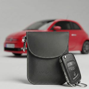 May include: A gray Faraday bag with a silver zipper and the text "FARADAY BAG" printed on the bottom. A black car key is next to the bag. A red car is in the background.