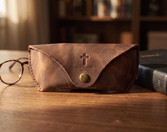 Könnte beinhalten: Ein braunes Brillenetui aus Leder mit dreieckigem Klappverschluss, der durch einen messingfarbenen Druckknopf gesichert ist. Auf der Vorderseite sind ein Kreuzsymbol und der Text "GOD IS LOVE JOHN 3:16" eingeprägt. Sichtbare Nähte an den Kanten.