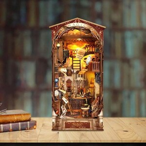 May include: A vertical book nook diorama featuring a detailed interior scene with warm interior lighting. The structure has an arched top with the word MAGIC and a base labeled MAGIC WORLD. It contains miniature furniture, books, and a broom.