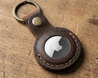 May include: A dark brown leather keychain holder featuring a circular cutout that secures a silver and black Apple AirTag. The leather is stitched with thick thread and includes a metal snap button and a circular metal key ring attached.