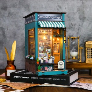 May include: A miniature wooden bookstore model titled Soul Bookstore with teal walls, a striped awning, and illuminated interior shelves filled with tiny books. The structure features a brick base, a sign reading Bookstore, and a small white sign with a book icon.