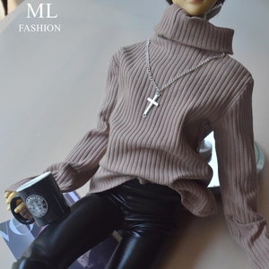 May include: A doll wearing a tan ribbed turtleneck sweater, black faux leather pants, and a silver cross necklace. The doll is holding a black mug with a white logo. The image has the text "ML FASHION" in the upper left corner.