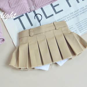 May include: A tan pleated skirt hangs on a black hanger. The skirt has a classic design with a waistband and pleats. A light pink jacket is partially visible in the upper left corner. The word "Moonlight" is written in cursive.