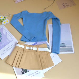 Puede incluir: Una blusa azul claro de manga larga y una falda plisada color beige con cinturón blanco y hebilla plateada. Las prendas se exhiben sobre una superficie de color beige claro con tarjetas decorativas y flores.