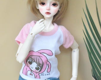 May include: A white doll with blonde hair wearing a pink and white t-shirt with a cartoon bunny design and blue denim shorts with white lace trim. The doll is wearing white cat ears.