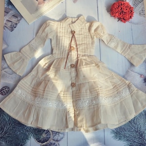 May include: A cream-colored dress with a high collar, long sleeves, and a tiered skirt. The dress features decorative buttons, lace trim, and a brown tie closure. The dress is made of a lightweight fabric.