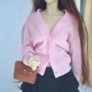 May include: A doll dressed in a light pink cardigan with three buttons and a black pleated skirt, holding a small brown purse. The word "Moonlig" is visible in the upper right corner. The cardigan has two pockets.