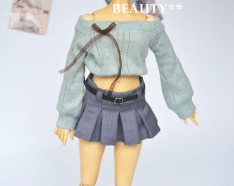 Puede incluir: Una muñeca con un suéter azul claro con hombros descubiertos con una cinta marrón y una falda plisada gris. La falda tiene un cinturón negro con una hebilla plateada. La palabra "BEAUTY**" está arriba.