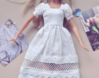May include: A white doll wearing a white dress with lace trim. The dress has a square neckline and short sleeves. The doll has blonde hair and blue eyes. The doll is wearing a white dress with lace trim. The dress has a square neckline and short sleeves. The doll has blonde hair and blue eyes.