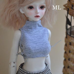 May include: A doll with pale skin, wearing a gray crop top with lace-up detailing and a black and white houndstooth skirt. The doll has light blonde hair and is posed in front of a neutral background. The letters "ML" are visible.