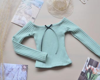 May include: A light green off-the-shoulder long-sleeved shirt with a black ribbon tied at the neckline.