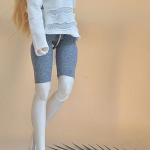 以下が含まれることがあります： 白いダメージ加工の長袖シャツとグレーのカプリレギンスを着た人形。人形は長い金髪で、金属製のスタンドに立っています。「Moonlight」の文字が右上部に筆記体で書かれています。