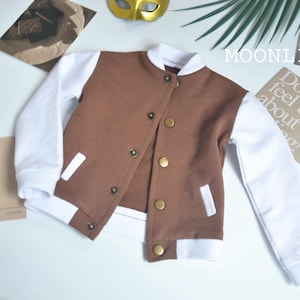May include: A brown and white varsity jacket with gold buttons. The jacket has a brown body and white sleeves, cuffs, and collar. The jacket is buttoned up and has white pocket accents. The word "MOONLIGHT" is visible in the image.