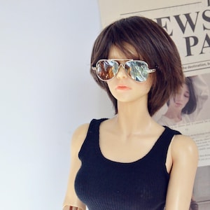 Puede incluir: Una mujer con el pelo corto y castaño que lleva un top negro de tirantes y gafas de sol de aviador. Está de pie frente a un periódico con la palabra "PARÍS" impresa en él.