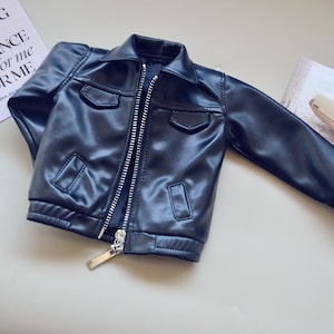 May include: A black faux leather jacket with a silver zip, two chest pockets, and a collared neckline. The jacket is on a light surface next to a card with text.
