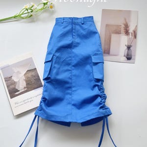 May include: A bright blue skirt with cargo pockets and drawstring details. The skirt has a high waist and a knee-length hemline. The word "Moonlight" is written in cursive at the top. The skirt is displayed on a white surface.