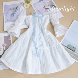 May include: A white off-the-shoulder dress with ruffled sleeves and a tiered skirt. A light blue ribbon is tied in a bow at the neckline. The word "Moonlight" is printed in white on a magazine.