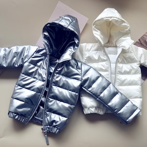 May include: Two hooded puffer jackets, one silver and one white, are displayed. Both jackets have a shiny, quilted texture, full zippers, and matching hoods. The jackets are laid out on a neutral surface, showcasing their design and style.