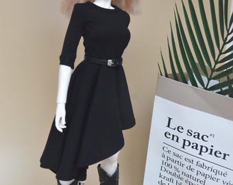 May include: A doll with blonde hair wearing a black dress and black boots. The doll is standing on a white platform. The background is a light brown wall with a green plant and a white sign with the text "Le sac en papier" in black.