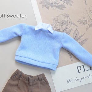 May include: A pale blue sweater with a white collar, alongside brown trousers. The words "Soft Sweater" are above the sweater. The outfit is displayed on a floral background.