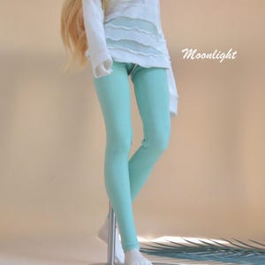 以下が含まれることがあります： テクスチャードデザインの白い長袖シャツと水色のレギンスを着た人形。 「Moonlight」の文字が画像の右側に筆記体で書かれています。