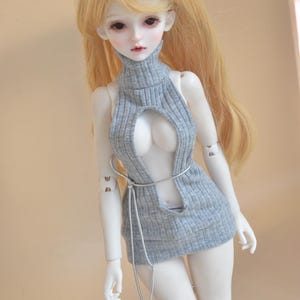 May include: A doll with long blonde hair wearing a gray, ribbed, open-front sweater dress. The dress has a high neck and a silver-colored tie around the waist. The word "Moonlight" is written in cursive at the top.