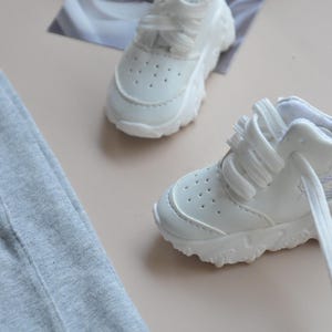May include: A pair of white sneakers with white laces. The sneakers have a chunky sole and are made of a smooth, white leather.