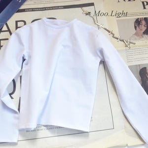 Op de afbeelding: Een witte longsleeve met een ronde hals, liggend op een krant. De shirt is gemaakt van een gladde stof. De krant heeft tekst en afbeeldingen, met de woorden "PARIS" en "Moo.Light" zichtbaar.