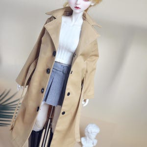 Peut inclure: Une poupée vêtue d'un trench-coat beige, d'un pull blanc et d'une jupe plissée grise. La poupée a les cheveux blonds et des bottes noires. Le texte "Moo.Light" est dans le coin supérieur gauche. Une petite statue de buste blanc est en arrière-plan.