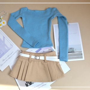 May include: A pale blue long-sleeved top and a beige pleated skirt with a white belt. The outfit is displayed on a light brown surface with decorative cards and flowers.