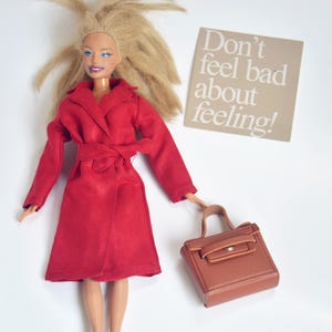 May include: A doll in a red coat and holding a brown handbag. The doll has blonde hair and is wearing a red coat with a belt. A sign that says "Don't feel bad about feeling!" is in the background.
