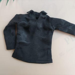 May include: A black long-sleeved shirt with a V-neck collar. The shirt has a subtle floral pattern and is made of a smooth, possibly satin-like fabric. The garment is laid flat, showcasing its design.