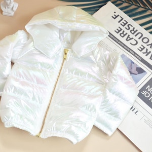 May include: A white, iridescent puffer jacket with a hood and a gold zip. The jacket is on a light beige surface next to a magazine and a newspaper with the text "TAKE CARE OF YOURSELF."
