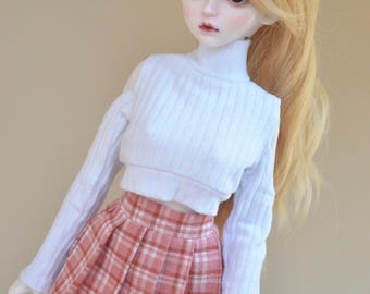 Puede incluir: Una muñeca con un suéter blanco de cuello alto de manga larga y una falda de cuadros rosa y blanco. La muñeca tiene el pelo largo y rubio.