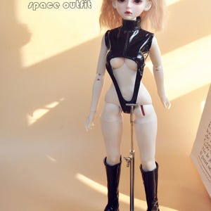 May include: A black vinyl doll outfit with a high neck and a cut-out design. The outfit is worn by a doll with blonde hair and black boots.