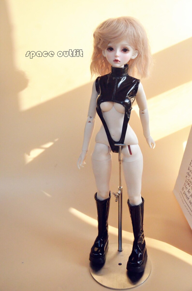 May include: A black vinyl doll outfit with a high neck and a cut-out design. The outfit is worn by a doll with blonde hair and black boots.