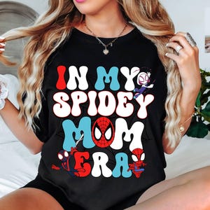 May include: Black t-shirt with the text 'In My Spidey Mom Era' in colorful retro font. The shirt features a spider-man design and other spider-man characters.