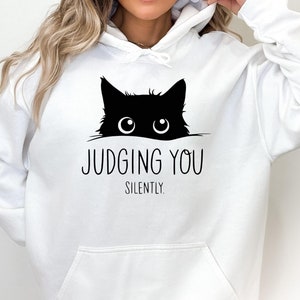 May include: White hoodie with a black cat peeking out from the top and the text "JUDGING YOU SILENTLY".