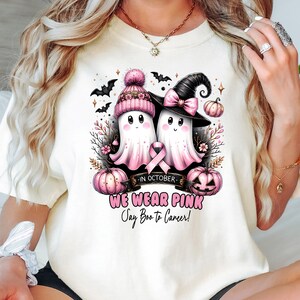 Op de afbeelding: Cr&egrave;mekleurig T-shirt met een afbeelding van twee cartoongeesten met roze hoeden en een roze lint, met pompoenen en vleermuizen. De tekst "WE WEAR PINK" en "Say Boo to Cancer!" wordt ook weergegeven.
