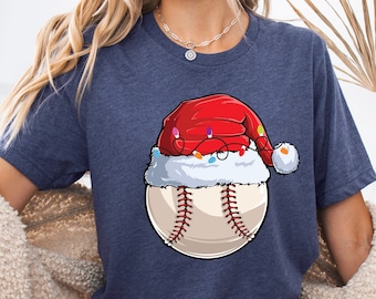 May include: Navy blue t-shirt featuring a baseball with a Santa hat and string lights. The baseball has red stitching and a white background. The Santa hat is red with white trim and a white pom-pom.