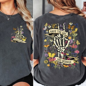 K&ouml;nnte beinhalten: Ein dunkelgraues T-Shirt mit einem Skelett-Hand-Motiv und dem Text "Have the day you deserve" auf der R&uuml;ckseite. Die Vorderseite des Shirts hat eine kleinere Version des Motivs mit dem Text "Have the day you deserve".