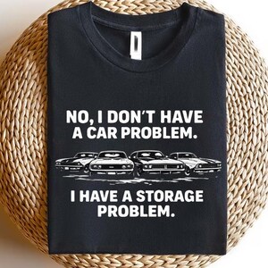 May include: Black t-shirt with the text "NO, I DON'T HAVE A CAR PROBLEM. I HAVE A STORAGE PROBLEM." and a graphic of four classic cars in white. The shirt is displayed on a woven, circular surface.