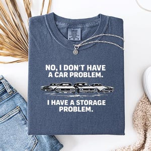 May include: A navy blue t-shirt with the text "NO, I DON'T HAVE A CAR PROBLEM. I HAVE A STORAGE PROBLEM." and a graphic of four classic cars. The shirt is laid flat with a pair of blue jeans, white shoes, and a necklace.