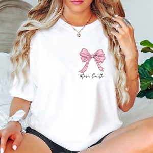 Op de afbeelding: Wit T-shirt met een roze strik en de tekst "Mrs. Smith" gedrukt op de voorkant.