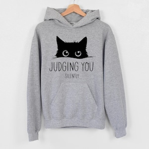 May include: A gray hooded sweatshirt with a black cat peeking out from the top and the text "JUDGING YOU SILENTLY".