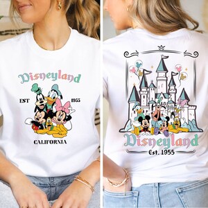 May include: White t-shirt with a colorful Disneyland graphic. The front of the shirt features Mickey Mouse, Minnie Mouse, Goofy, Donald Duck, and Pluto. The back of the shirt features a colorful illustration of Sleeping Beauty's castle with the text "Disneyland Est. 1955".