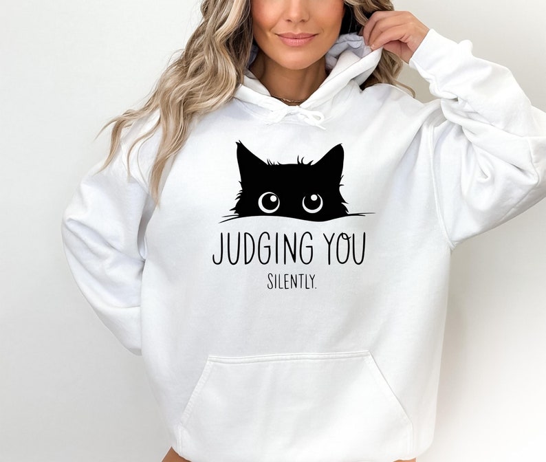 May include: White hoodie with a black cat peeking out from the top and the text "JUDGING YOU SILENTLY".