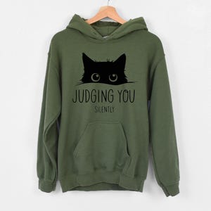 May include: A green hooded sweatshirt with a black cat peeking out from behind the text "JUDGING YOU SILENTLY".